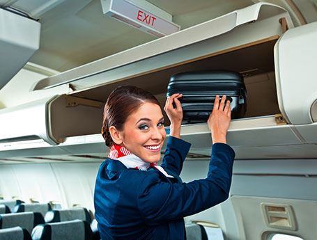 Female flight attendant places a small suitcase in an overhead bin while smiling at the camera