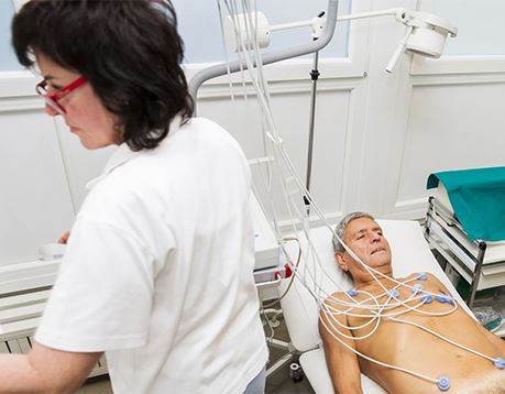 A cardiovascular technician stands near a patient who has been prepped with electrodes for an EKG