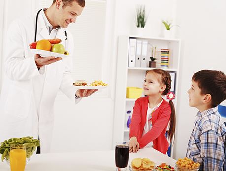 Nutritionist smiles and holds a plate of fruit and a plate of burgers in front of two young children