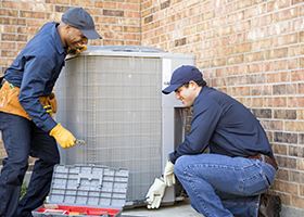 Two HVACR workers inspect an air-conditioning unit outside of a building
