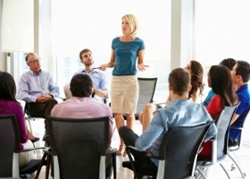 Business woman speaks standing in the middle of a circle of other business people sitting in chairs