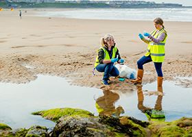 Two female marine scientists collect water samples from a shallow pool of water on the beach