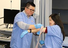 A personal care aide student wraps the arms of another student in a simulated hospital environment