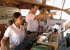 Three professional mariners navigate a ship while one looks through binoculars
