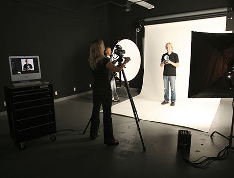 Woman takes a photograph of a man against a white backdrop in a photo studio