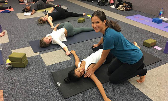 Female yoga instructor smiles at camera as she corrects form of student