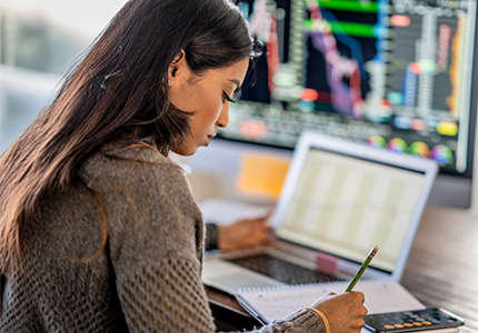 A woman taking notes while analyzin data on her laptop.