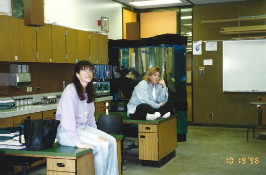 Two students sitting on top of desks in a classroom with an aquarium behind them at the front of the classroom, 1996.