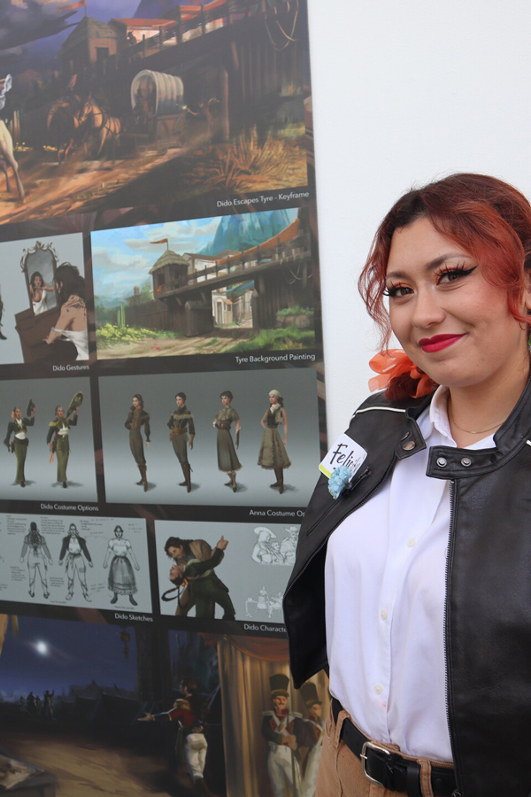 A woman with red hair smiles confidently in front of a display showcasing character designs and background paintings.