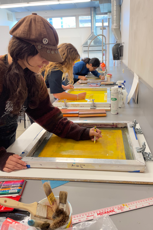 A student working on a screen printing project at a table surrounded by supplies and classmates.