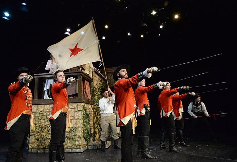 Actors onstage in red uniforms salute with swords
