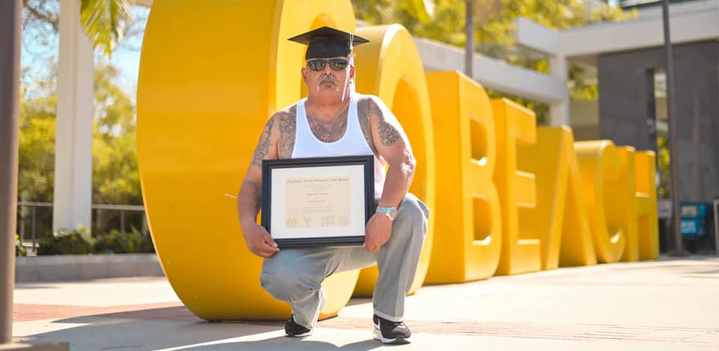 photo of kneeling man holding degree in front of large letters spelling out words go beach