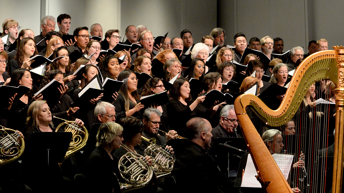 photograph of orchestra members dressed in black. In the top half of the photo are singers holding sheet music. In the bottom half of the photo are instrumentalists playing french horns, trumpets, clarinets and a large harp. 