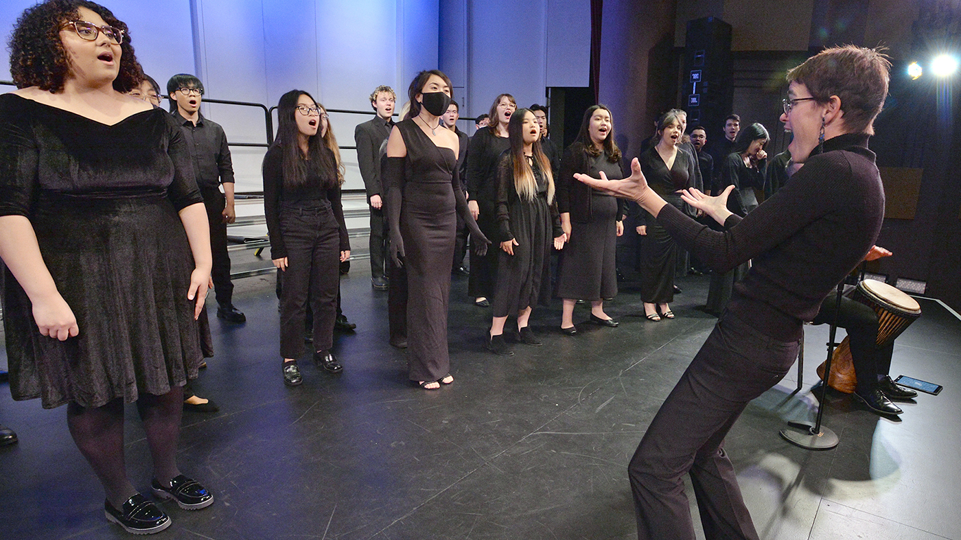 A choir group, dressed in black, appears to be singing on stage with one member conducting them enthusiastically.