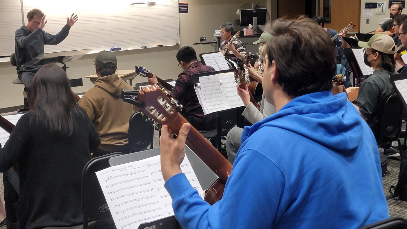 A group of adult students sitting in a classroom setting, playing guitars while a conductor gestures energetically at the front.