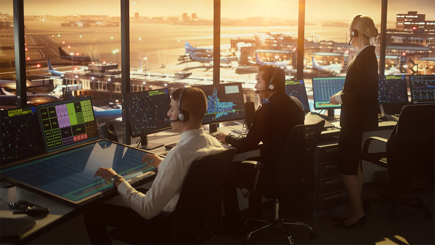 Air traffic control room with three individuals wearing headsets, seated at consoles with multiple screens displaying radar and flight data. Large windows reveal a busy airport runway and several aircraft, with a sunset in the background.