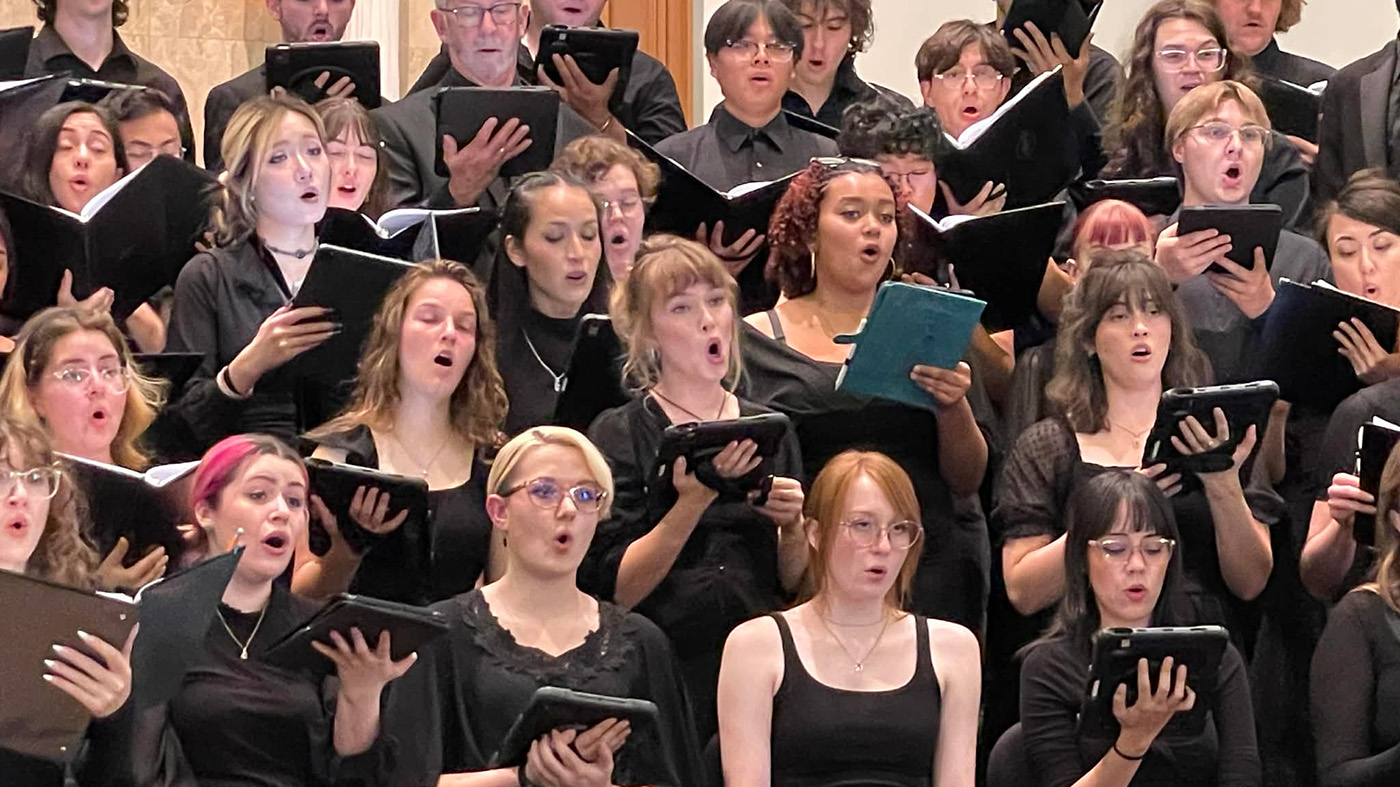 A large choir dressed in black performs indoors, holding music folders.