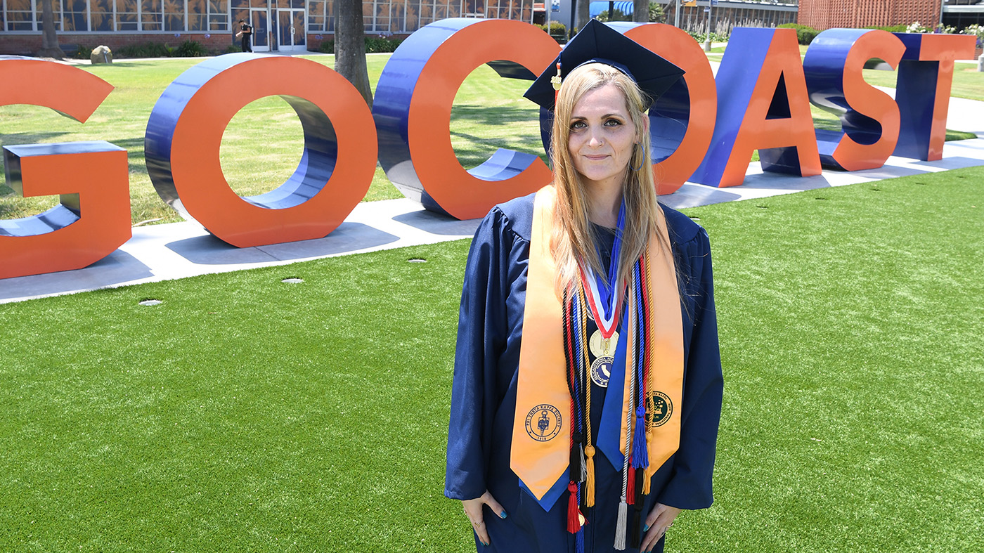 A woman in a graduation gown stands on a green lawn with large orange "GO COAST" letters in the background.