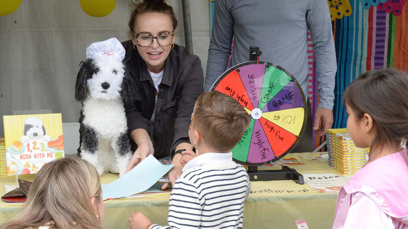 A person interacts with children at a booth featuring a prize wheel and a fluffy dog puppet wearing a chef's hat.