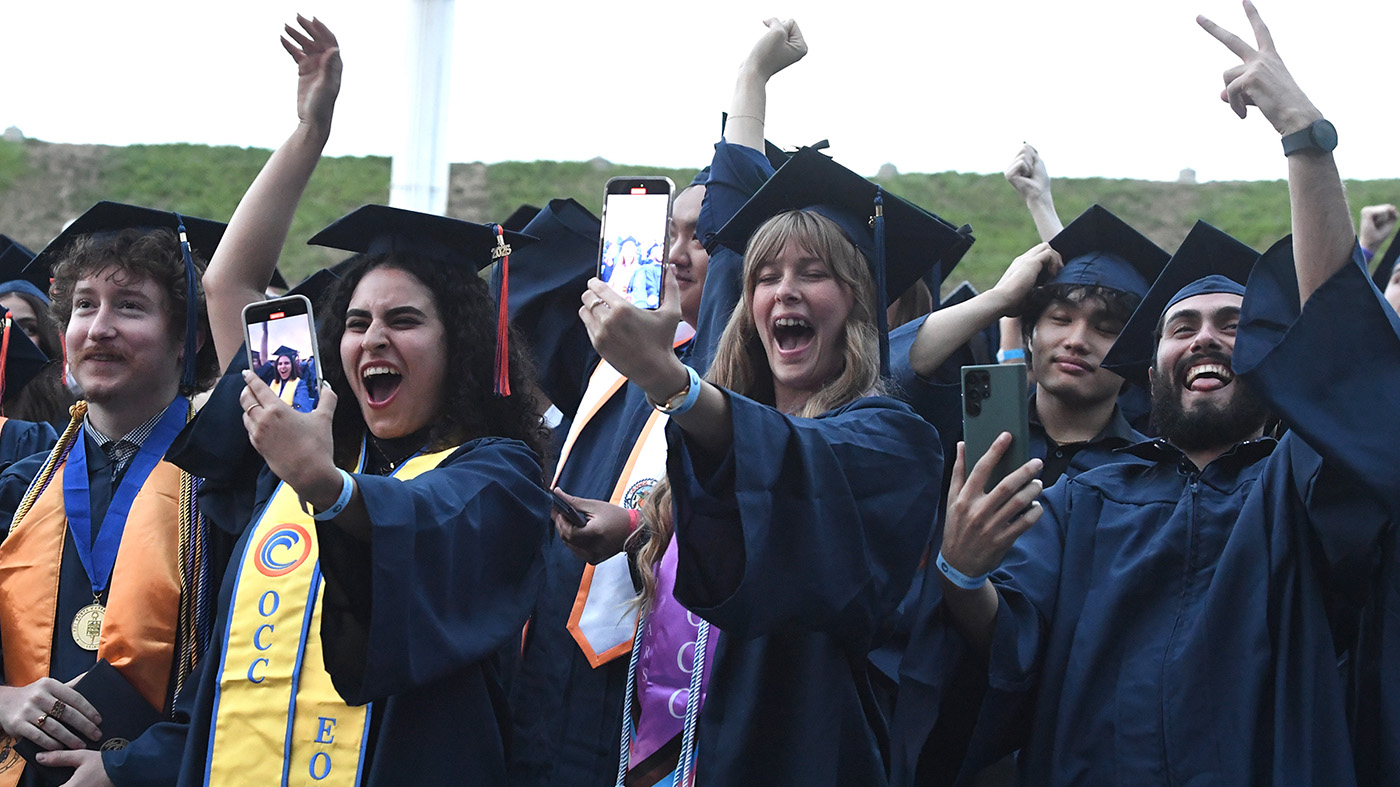 Graduates in navy gowns celebrate with smartphones held up, smiling and cheering outdoors.