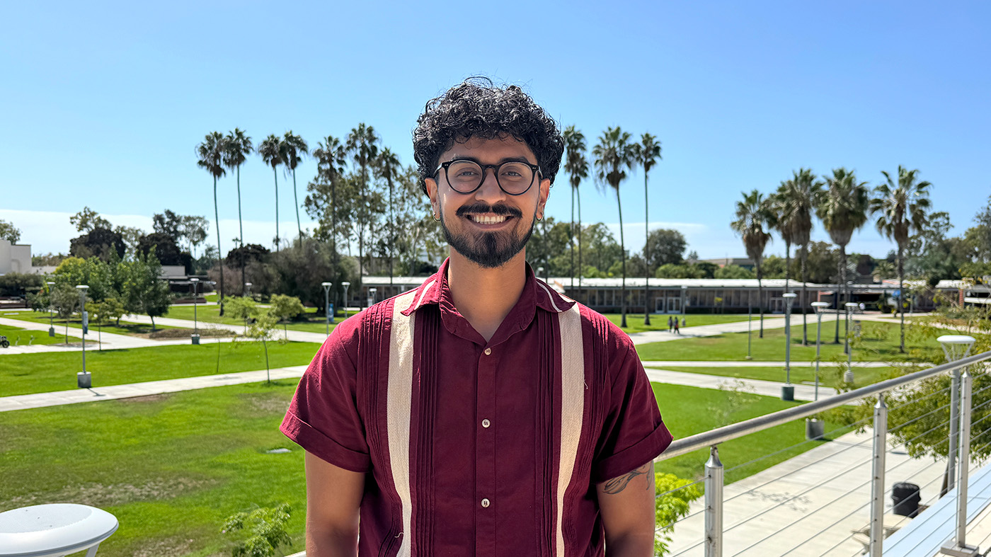 A person is smiling while standing on a balcony overlooking a green lawn. The person is wearing glasses and a red short-sleeved shirt. 