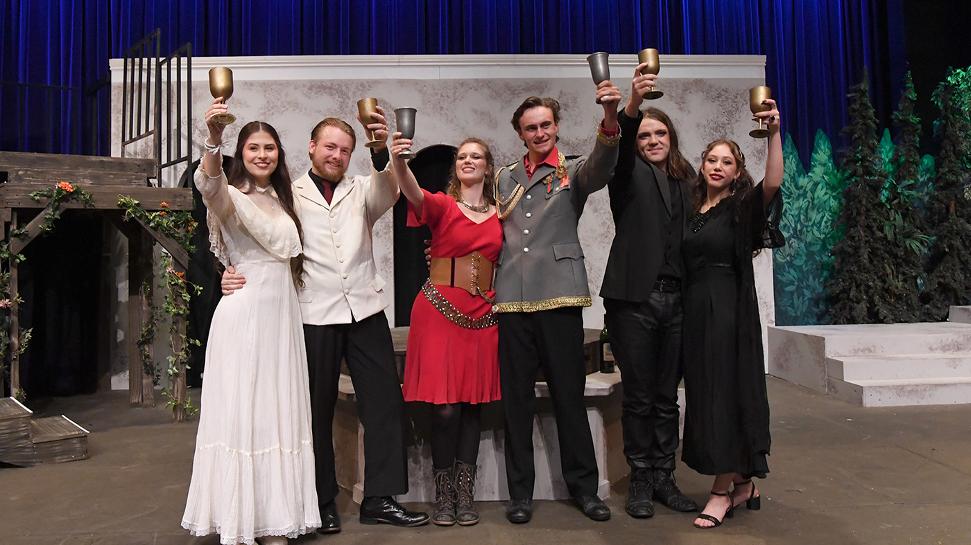 A group of six people standing on a stage, all holding goblets raised in a toast. They are dressed in various costumes, including formal and historical attire. The backdrop features a set with textured walls and a blue curtain.