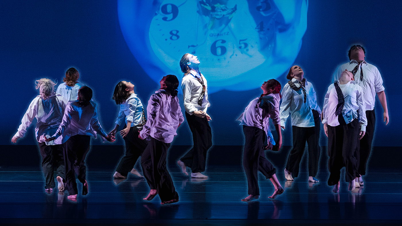 A group of dancers in expressive poses on a dimly lit stage with a clock projected in the background.