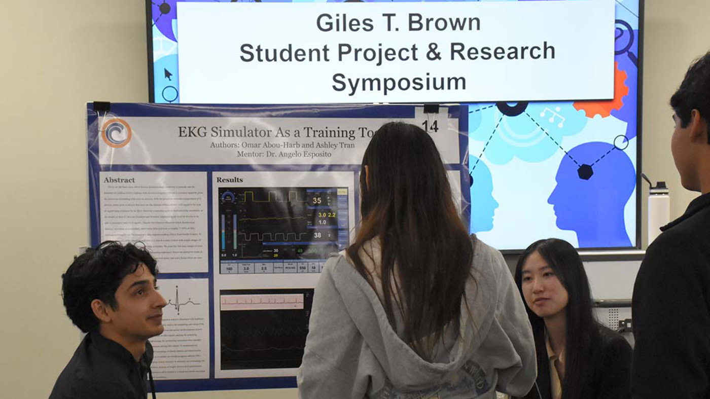 Students presenting an EKG Simulator project at a research symposium with a visible digital sign in the background.