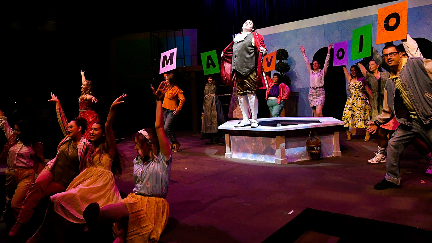 A stage performance with performers in colorful attire, some holding large cards. A spotlight highlights a person on a platform.