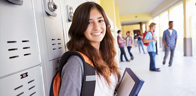A student with a backpack holding books near lockers in a hallway.