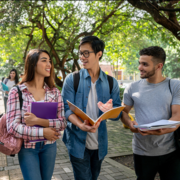 3 student walking together on campus holding notebooks.